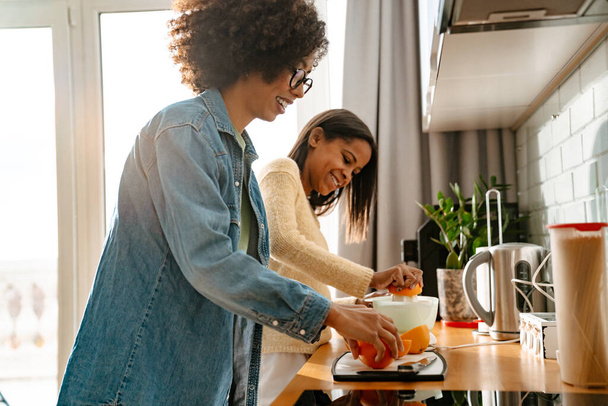 Happy african american woman and her teenage daughter making fresh orange juice in kitchen - Photo, Image