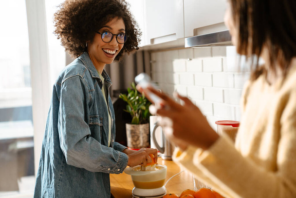 Black girl using mobile phone while talking with her mother in kitchen at home - Foto, Imagen