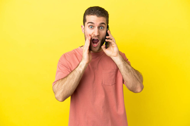 Όμορφος ξανθός άντρας που χρησιμοποιεί κινητό τηλέφωνο σε απομονωμένο φόντο με έκπληξη και σοκαρισμένη έκφραση προσώπου - Φωτογραφία, εικόνα