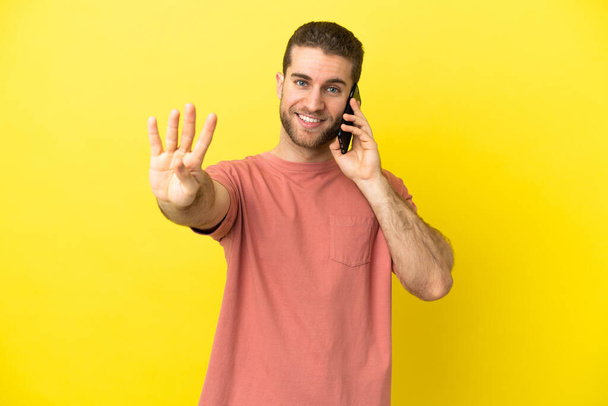 Yakışıklı sarışın adam izole edilmiş arka planda cep telefonu kullanıyor mutlu ve parmakları olan dört kişi sayıyor. - Fotoğraf, Görsel