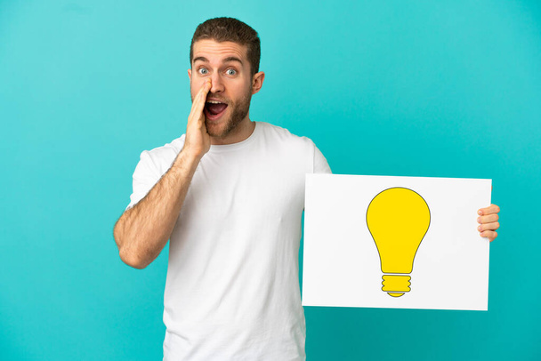 Handsome blonde man over isolated blue background holding a placard with bulb icon and shouting - Foto, Imagen