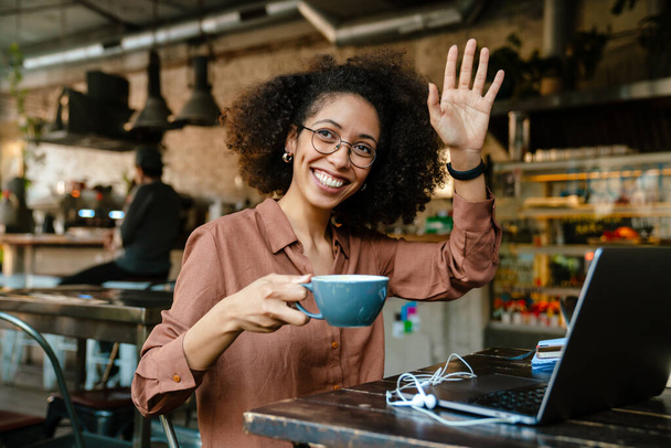 Young african american woman waving and drinking coffee while sitting by table in cafe - Foto, afbeelding