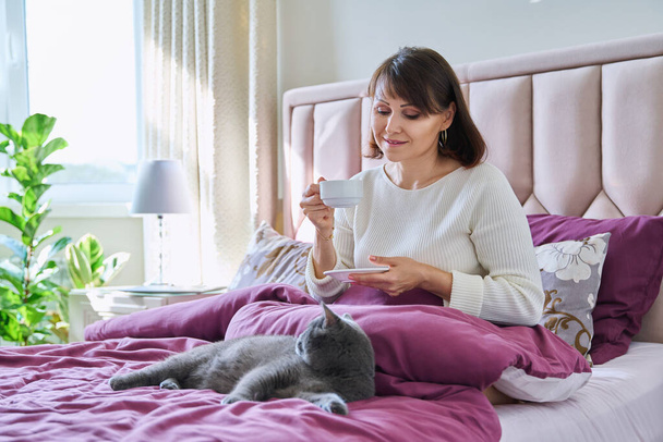 Middle aged beautiful relaxed woman drinking coffee in bed. Mature female holding cup of coffee, owner and pet grey cat lying together in morning in bedroom - Фото, изображение