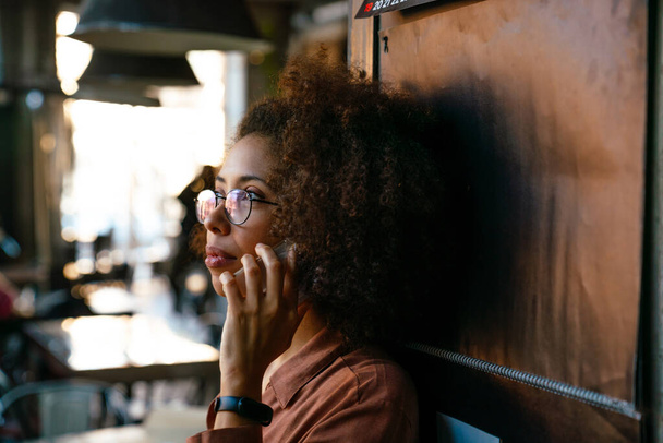 Young african american woman wearing eyeglasses talking on cellphone in cafe - Fotó, kép