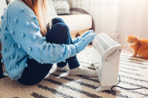 Using heater at home in cold winter. Woman warming her hands sitting by device with cat and wearing sweater and warm clothes. Heating season. - Photo, Image