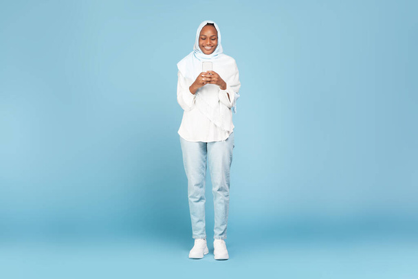 Полнометражный снимок счастливой чернокожей мусульманки, использующей современный смартфон на синем фоне студии, проверяющей новейшее развлекательное мобильное приложение, копирующее пространство - Фото, изображение