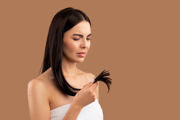 Unhappy half-naked brunette young woman holding her long hair split ends in hand, isolated on beige background, need for haircare mask treatment, diet correction, balm, vitamins, copy space for advert - Photo, Image