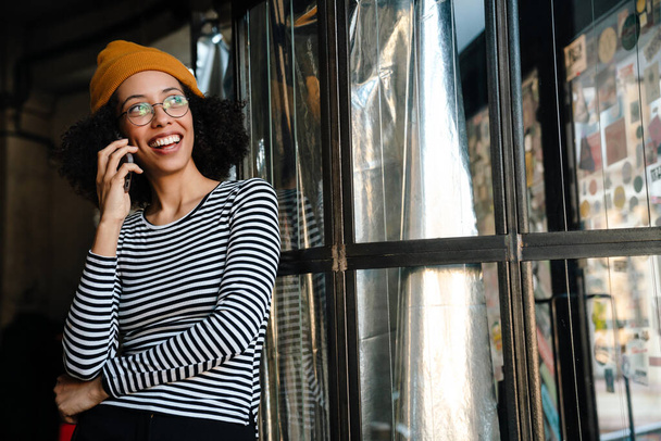 Joven mujer afroamericana sonriendo mientras habla por teléfono celular en la cafetería - Foto, Imagen
