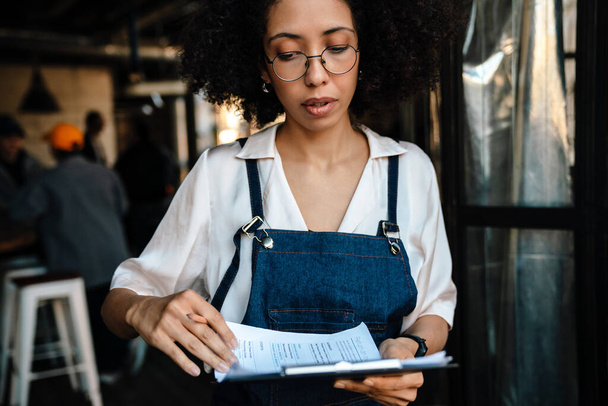 Young african american woman holding clipboard with paper documents while working in cafe - Foto, Imagen