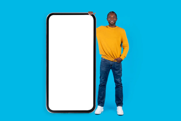 陽気クール大人黒男でカジュアルポージングで巨大なスマートフォンで白い空の画面上の青いスタジオの背景に,経由して女性とチャット日付アプリ,素晴らしいオファーを表示,モックアップ,フル長さ - 写真・画像