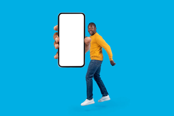 Suuri online tarjous. Innoissaan iloinen keski-ikäinen afrikkalainen amerikkalainen mies tyylikäs rento asu hyppäämällä ilmassa moderni älypuhelin valkoinen tyhjä näyttö eristetty sininen, mockup, täyspitkä - Valokuva, kuva