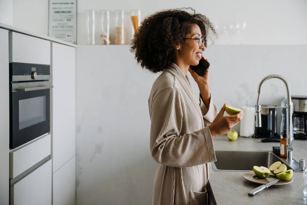 African american young woman with curly afro hairstyle using cellphone and eating apple in kitchen - Photo, Image
