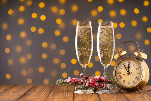 Two wine glasses with bubbly champagne, fir branch with decor and bronze clock on background of blurry sparkling lights. Happy New Year holiday greeting card, banner, header with copy space - Photo, Image