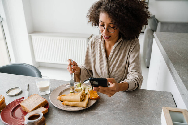 Black woman wearing eyeglasses using mobile phone while having breakfast at home - Fotó, kép