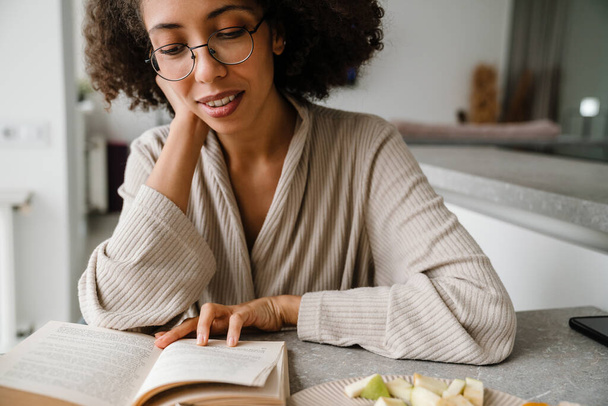Black woman wearing eyeglasses reading book while having breakfast at home - Fotó, kép