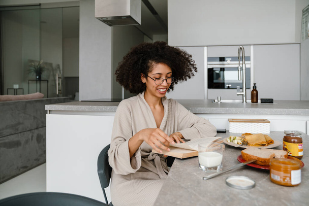 Black woman wearing eyeglasses reading book while having breakfast at home - Foto, Imagem