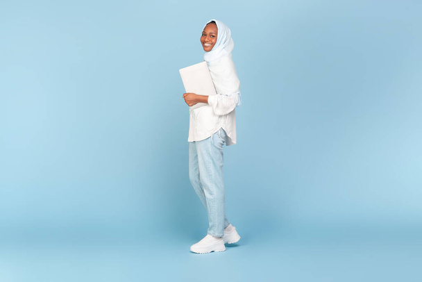 Happy african american woman in hijab holding laptop and walking on blue studio background, full length shot, free space. Internet business and freelance career concept - Photo, Image