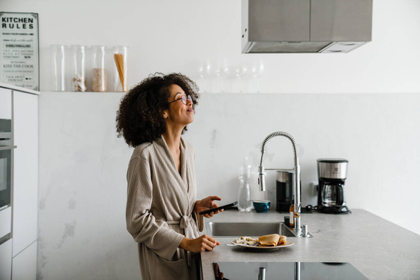 Black woman wearing eyeglasses using mobile phone while having breakfast at home - Foto, Imagen