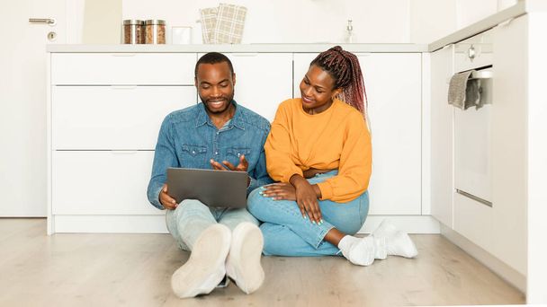 Excited african american spouses relaxing with laptop while sitting on floor in kitchen, shopping online or browsing internet on computer at home, panorama, copy space - Фото, зображення
