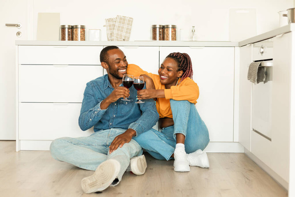 Ρομαντικό ζευγάρι Αφροαμερικανών που πίνουν κόκκινο κρασί ενώ κάθονται στο πάτωμα της κουζίνας, σύζυγοι που κρατούν γυαλιά και χαμογελούν ο ένας στον άλλο, γιορτάζοντας την επέτειο στο σπίτι - Φωτογραφία, εικόνα