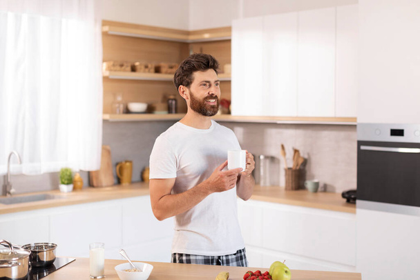 Χαρούμενος μεσήλικας καυκάσιος άνδρας με γενειάδα σε λευκό t-shirt ποτά ζεστό ρόφημα κοιτάζει κενό χώρο στο σύγχρονο εσωτερικό της κουζίνας. Διάλειμμα για καφέ στο σπίτι, καλημέρα και πρωινό, μεγάλη διαφήμιση και προσφορά - Φωτογραφία, εικόνα