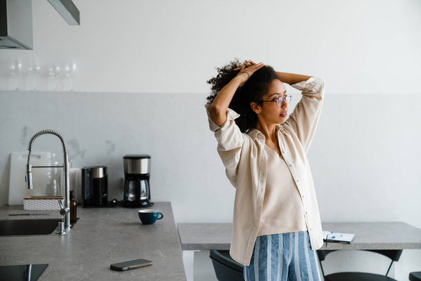 Black woman wearing eyeglasses holding her hair while standing in kitchen at home - Фото, зображення