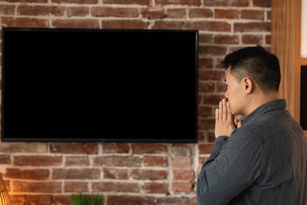 Печально обеспокоенный азиатский мужчина средних лет смотреть новости на большом телевизоре с пустым экраном в интерьере домашнего офиса, профиль. Мировой кризис, плохие новости, акции, инвестиции для бизнеса и работы во время пандемии ковид-19 - Фото, изображение
