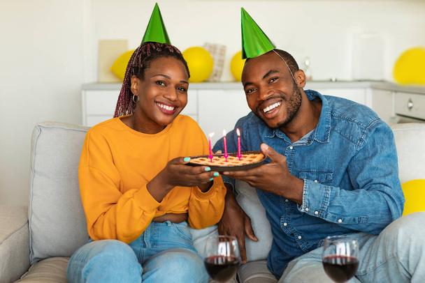 Портрет чёрной пары в шляпах для вечеринок, держащей пирог ручной работы с зажжёнными свечами и улыбающейся в камеру, сидящей дома на диване. Счастливые супруги празднуют день рождения вместе - Фото, изображение