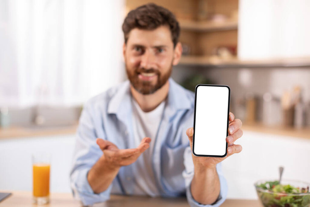 Portrait of glad adult caucasian man with beard shows phone with blank screen in kitchen interior. Recommendation and advice, app for work, business at social distance, device at home, offer and ad - Photo, image