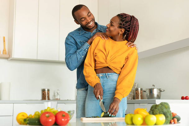 Szczęśliwy kochający czarni małżonkowie gotowanie zdrowego jedzenia razem, mąż obejmujący żonę z tyłu i uśmiechając się do siebie, przygotowując sałatkę warzywną w domu - Zdjęcie, obraz