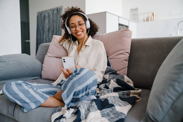 African american young woman using cellphone and headphones while sitting on sofa at home - Photo, Image