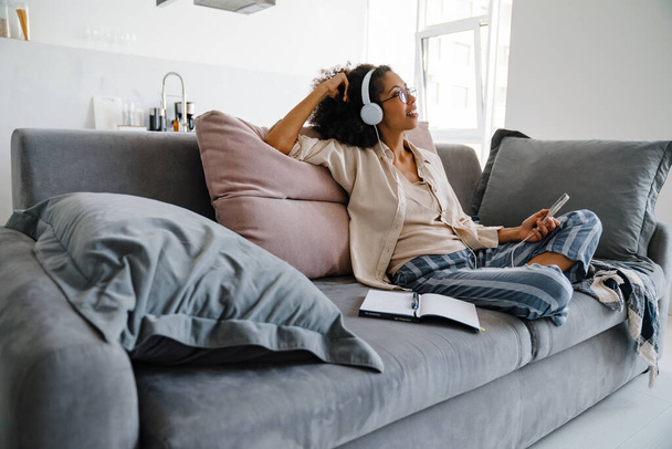 African american young woman using cellphone and headphones while sitting on sofa at home - Foto, afbeelding