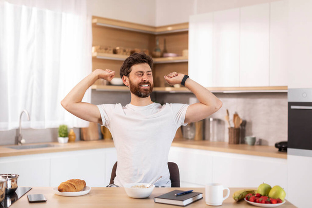 Glad cheerful adult caucasian man with beard in white t-shirt stretching his body in modern kitchen interior, free space. Guy enjoys good morning and breakfast at free time and weekend alone at home - Photo, Image