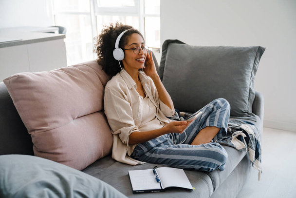 African american young woman using cellphone and headphones while sitting on sofa at home - Foto, immagini