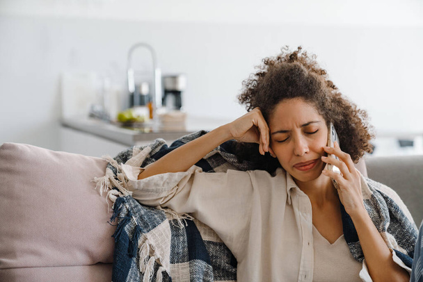 Черная недовольная женщина разговаривает по мобильному телефону, сидя дома на диване - Фото, изображение