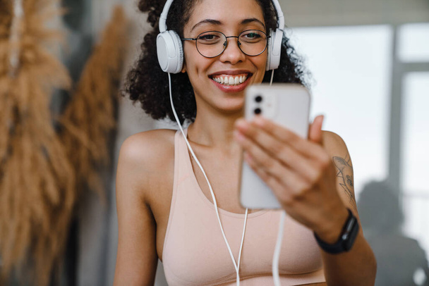 Black young woman wearing eyeglasses using cellphone and headphones during workout at home - Foto, Imagem