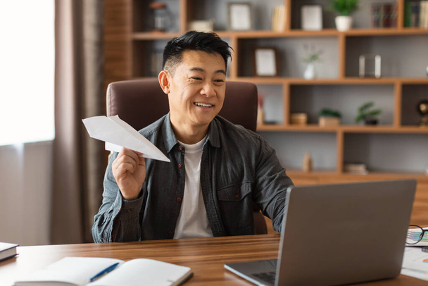 Улыбающийся азиатский мужчина средних лет делает самолет с бумагой, планируя стратегию за столом с ноутбуком в интерьере домашнего офиса. Создание идеи для бизнеса, брейншторм, работа во время пандемического covid-19 - Фото, изображение