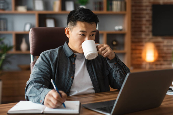 Зайнятий концентрований середнього віку азіатський чоловік п'є каву, дивиться на комп'ютер за столом, має відеодзвінок в інтер'єрі домашнього офісу. Відстань від роботи, бізнесу та фрілансу під час пандемії ковадла-19
 - Фото, зображення