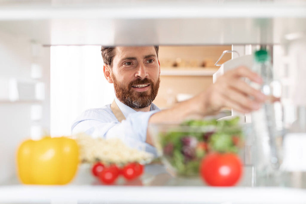 Lachende volwassen blanke man met baard in schort opent deur en kijkt naar koelkast met biologische groenten, neemt fles water in keuken interieur. Gezondheid en lichaamsverzorging, producten voor thuis koken - Foto, afbeelding