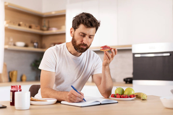 Busy concentrated adult caucasian male with beard in white t-shirt eats sandwich, makes notes in notebook in modern kitchen interior. Good morning and breakfast alone, study, planning and work at home - Photo, Image