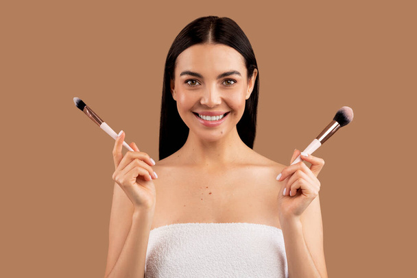 Photo of young happy smiling beautiful brunette half-naked woman holding makeup brushes, doing contouring or applying blush on face isolated on beige studio background - Foto, Imagem