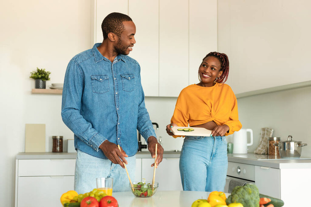 Emozionata coppia afro-americana che cucina insieme, preparando insalata di verdure fresche per la cena in cucina a casa. Ricette di famiglia, nutrizione sana e concetto di preparazione alimentare - Foto, immagini