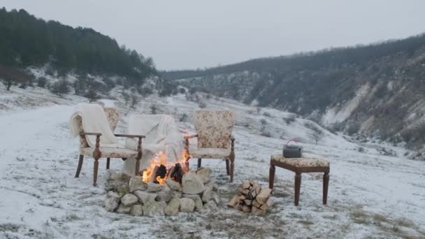 Пожар на снегу у горного ручья, в зимнем лесу с удобными креслами и пушистыми кроватями для прекрасной пары. Зимний пейзаж. Концепция приключений. - Кадры, видео