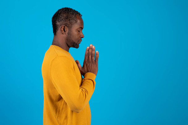 Boczny widok spokojnego duchowo przystojnego, dojrzałego czarnego mężczyzny modlącego się zamkniętymi oczami. Poważny, spokojny człowiek z połączonymi dłońmi medytujący. Odizolowany na tle niebieskiego studia. Koncepcja wierzeń, przestrzeń kopiowania - Zdjęcie, obraz