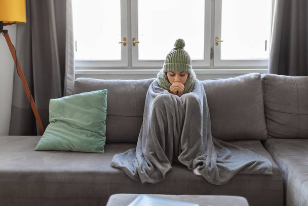 Jonge Arabische vrouw die thuis kou lijdt terwijl ze op de bank zit, bedekt met deken, duizendjarige vrouw met sjaal en gebreide muts die bevriest in de woonkamer, problemen heeft met het verwarmingssysteem - Foto, afbeelding