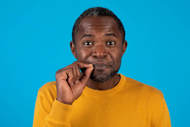 Schöner schwarzer Mann mittleren Alters in lässigem gelben Pullover, der Mund und Lippen als Reißverschluss mit Fingern geschlossen zeigt. Geheimhalten und Schweigen, Tabureden. Vereinzelt auf blauem Studiohintergrund - Foto, Bild