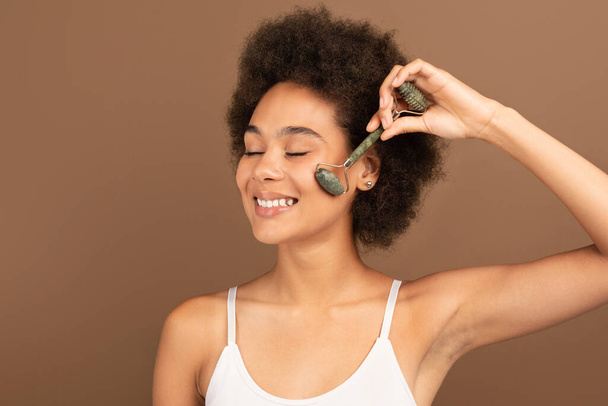 Sorrindo muito millennial afro-americano encaracolado fêmea em top branco aplicar blush ou pó no rosto com escova, isolado em fundo marrom, estúdio. Maquiagem nua, anúncios de cosméticos e cuidados de beleza em casa - Foto, Imagem