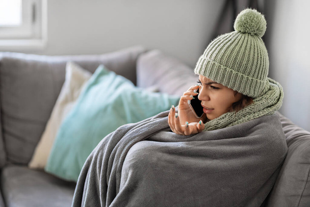 Jovem estressada conversando no celular enquanto sentada no sofá embrulhada em cobertor, inquilino fêmea perturbada congelando em casa, reclamando ao proprietário sobre baixa temperatura no quarto, vista lateral - Foto, Imagem