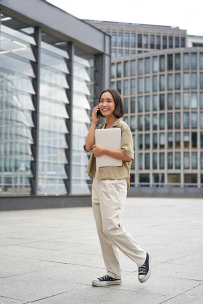 Vertical portrait of stylish asian girl student, talking on mobile phone while walking, holding laptop. - Zdjęcie, obraz