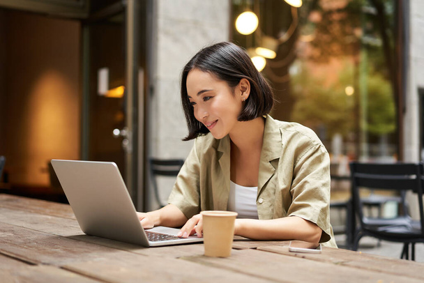 Joven mujer asiática, nómada digital trabajando remotamente desde un café, bebiendo café y utilizando el ordenador portátil, sonriendo. - Foto, Imagen